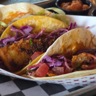 Crispy fish tacos filled with slaw, tomatoes, and sauce in a paper tray.