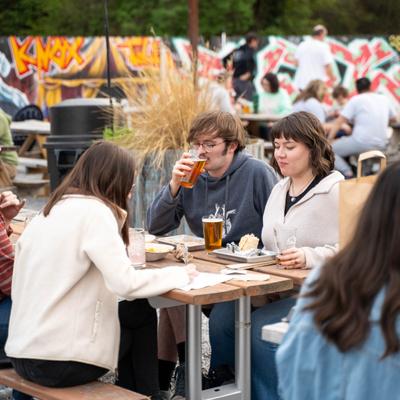 People seated at outdoor tables enjoying drinks with graffiti art on a wall in the background.