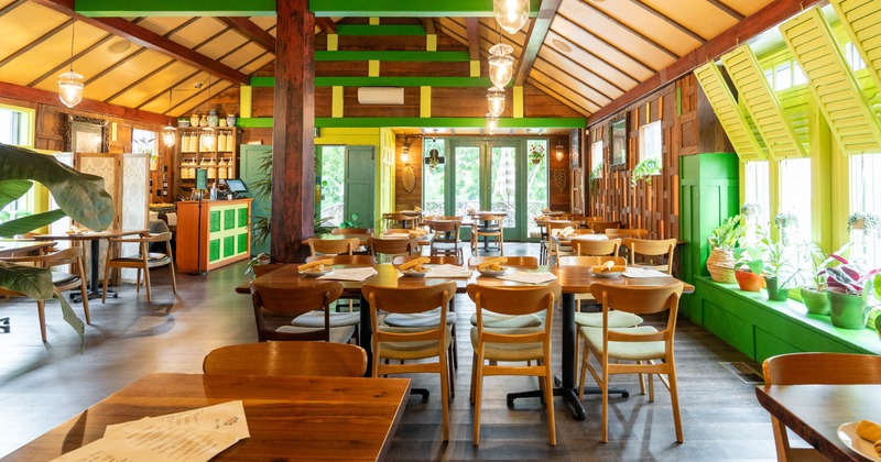 Interior with wooden tables and chairs, large windows with green shutters, and potted plants