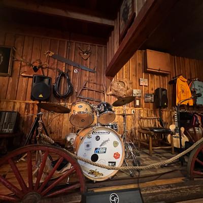 Drum kit on a rustic wooden stage surrounded by antlers, tools, and music equipment.
