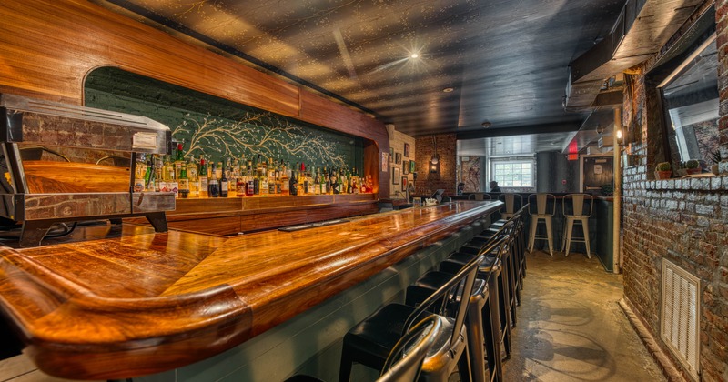 Bar interior with a long wooden counter, black metal stools and brick walls.