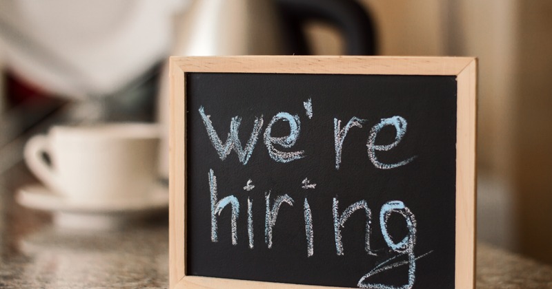 A chalkboard with the text We're hiring  written on it,  placed on a table with a coffee