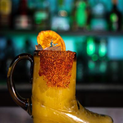 Cocktail served in chili-rimmed, boot-shaped glass with a dried orange wheel.