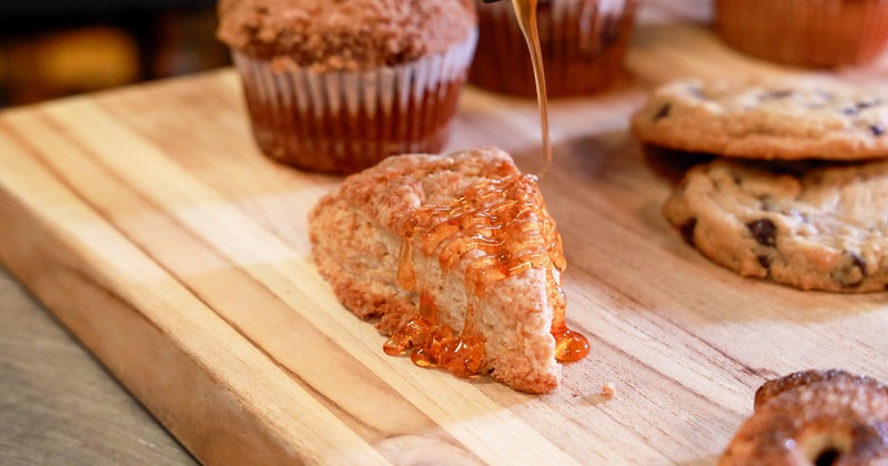Scone with honey drizzled on top sits on a wooden board surrounded by cookies and muffins