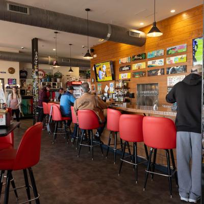 People sitting at a brewery bar with red stools and wall-mounted TVs.