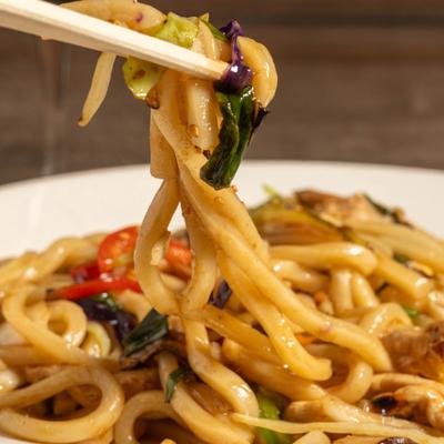 Chopsticks holding stir-fried udon noodles with vegetables, over the plate.