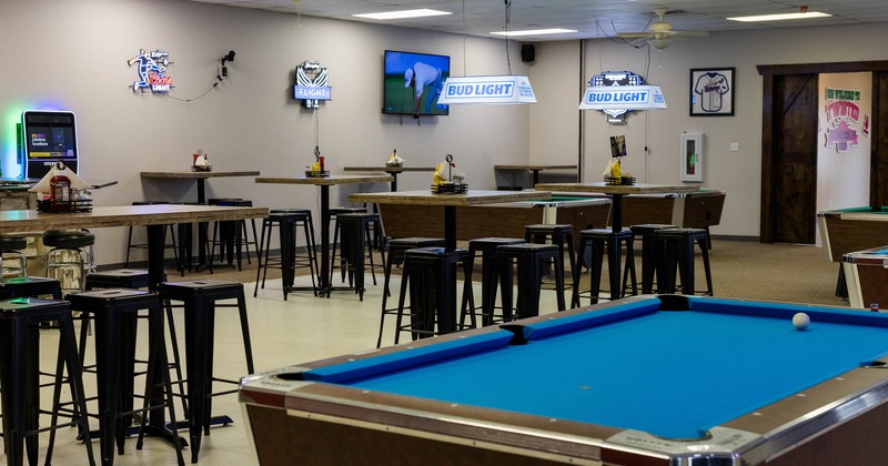 A bar area with high tables, stools, a pool table, neon beer signs and a TV