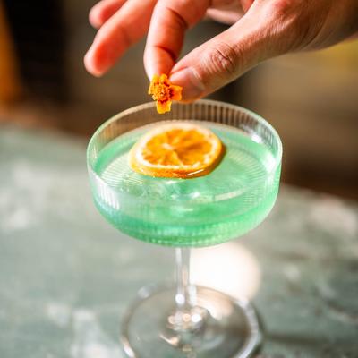 A light blue mocktail with a dry citrus slice, being garnished with an edible flower.