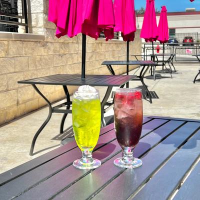 Two drinks on a table outside, tables adorned with umbrellas under the bright sun.