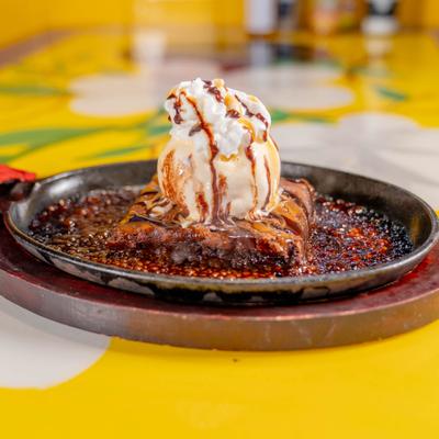 Sizzling brownie topped with vanilla ice cream and chocolate sauce on skillet.