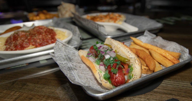 Chicago-style hot dog with fries on a tray, background shows pasta with sauce