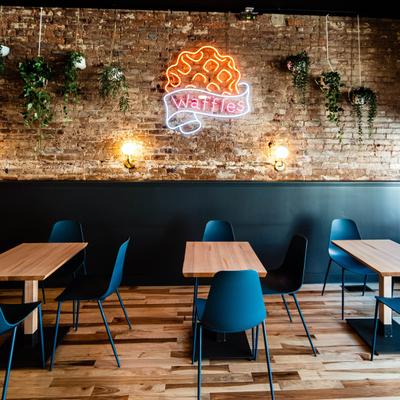 Interior with exposed brick walls, wooden tables, blue chairs and a neon sign.