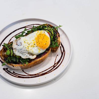 A slice of toasted bread topped with an egg, avocado, arugula, and balsamic glaze drizzle.