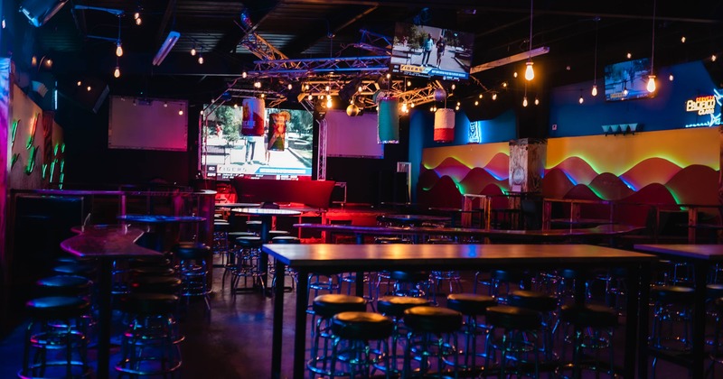Interior of a colorful bar with high tables, barstools, neon lights, and a large video screen