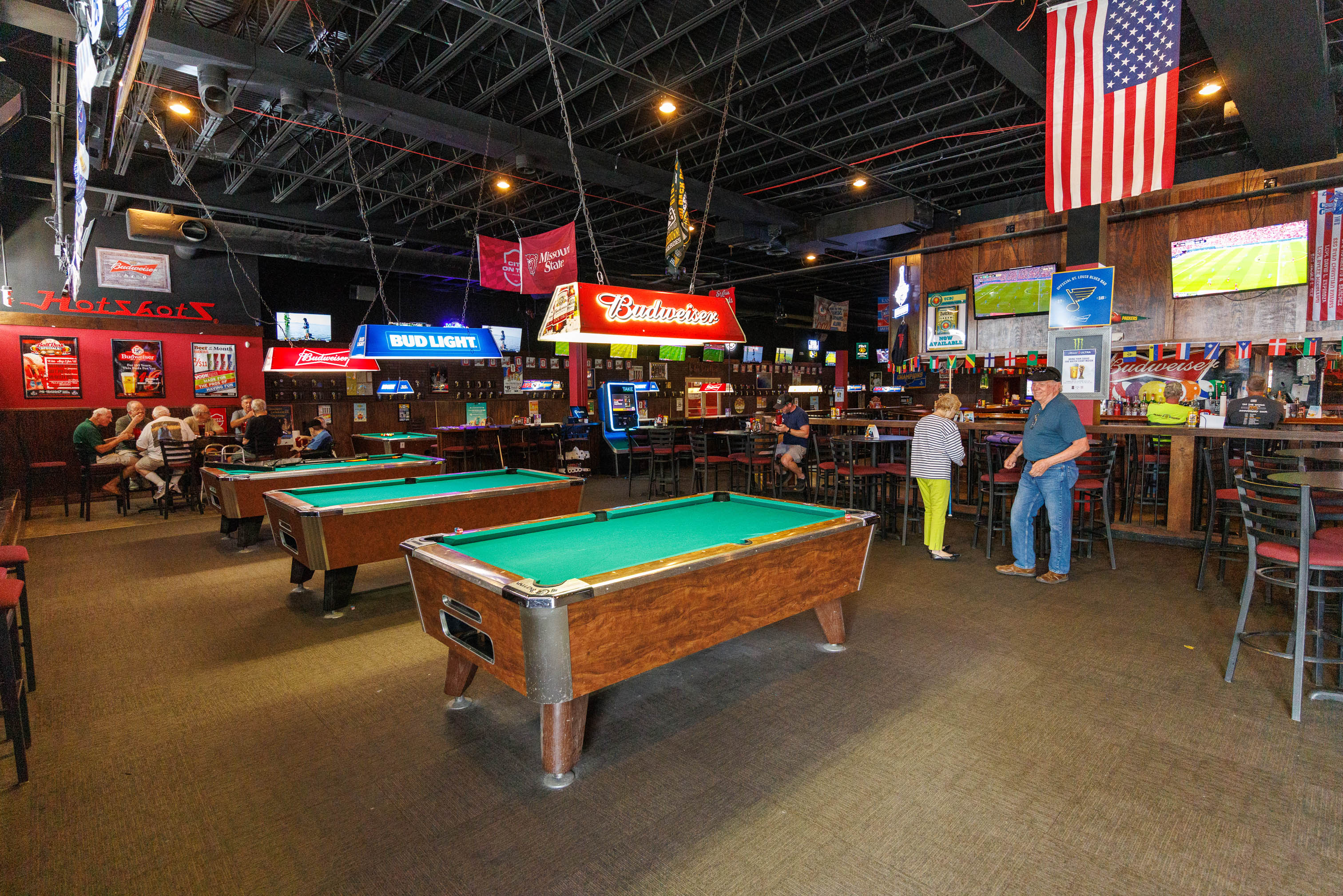 A spacious sports bar features multiple wooden pool tables under bright neon signs