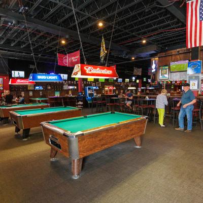A spacious sports bar features multiple wooden pool tables under bright neon signs.