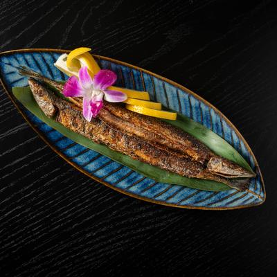 Grilled Pacific saury fish served on a plate with lemon slices and purple flower.