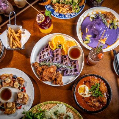 Colorful brunch spread with purple waffles, fried chicken, cocktails, and assorted dishes.