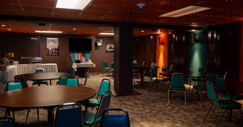 Indoor event space with round wooden tables, teal chairs, and a colorful wall design