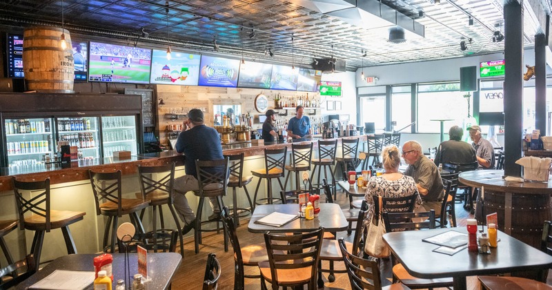 Patrons are seated at tables and the bar in the sports bar with multiple TVs displaying games