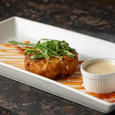 Blue crab cake topped with microgreens, served with a ramekin containing creamy sauce.