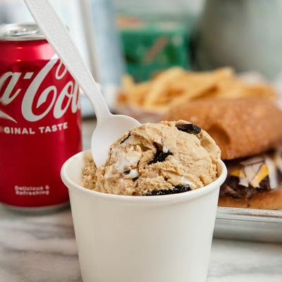 A paper cup holds a scoop of ice cream in front of canned soda and a burger plate.