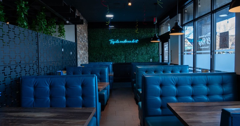 Interior of a restaurant with blue tufted booths, a greenery wall with neon sign, and large windows