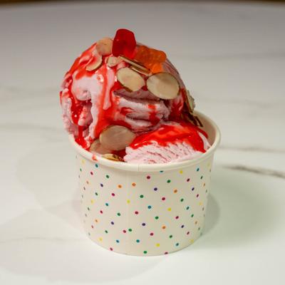 Cup of rose ice cream topped with sliced almonds, red sauce and gummy bears.