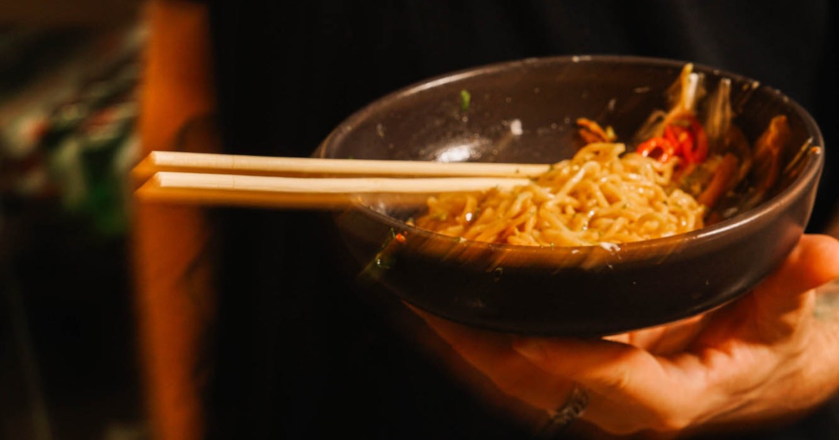 A hand holds a dark bowl of noodles with wooden chopsticks