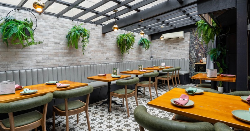A bright indoor dining space with hanging ferns, wooden tables, green chairs, and a glass ceiling