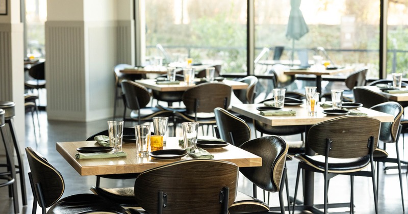 Restaurant interior with wooden tables set with plates, napkins, and glasses, large windows