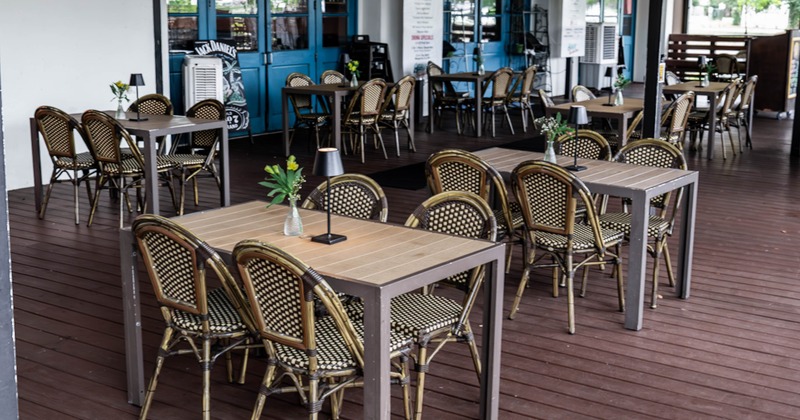 Outdoor restaurant patio with blue doors, wooden tables, woven chairs, and small floral decorations