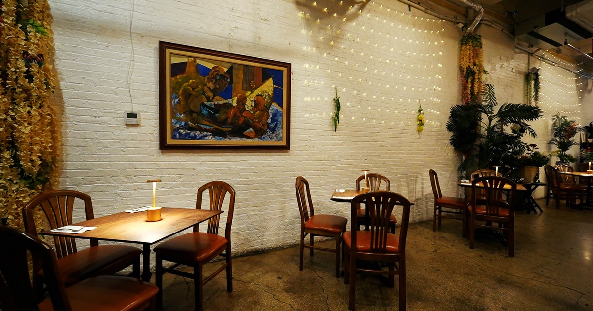 A cozy interior - white brick walls, a framed painting, wooden tables and chairs; warm lighting