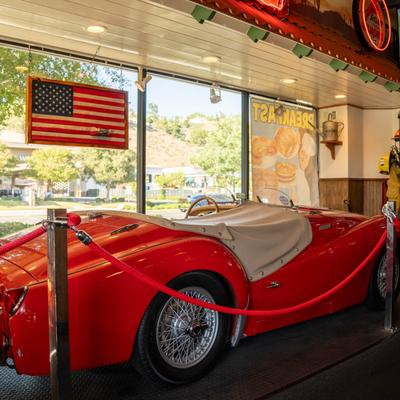 A vintage red convertible sports car is displayed indoors with a gas pump and American flag.