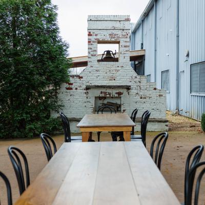 Outdoor patio seating with long wooden tables, black chairs, and rustic brick fireplace.