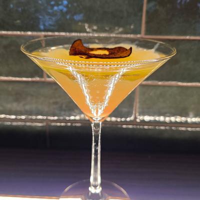 Yellow cocktail in a martini glass garnished with a dark, dried fruit slice.