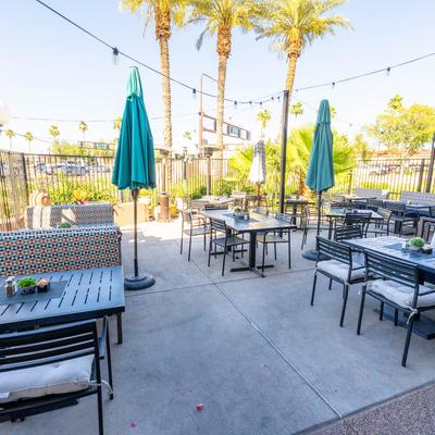Outdoor patio with black tables, chairs, and parasols set against palm trees and clear sky.