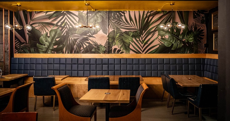 A dining area with a tropical leaf mural, navy blue banquette and wooden tables and chairs