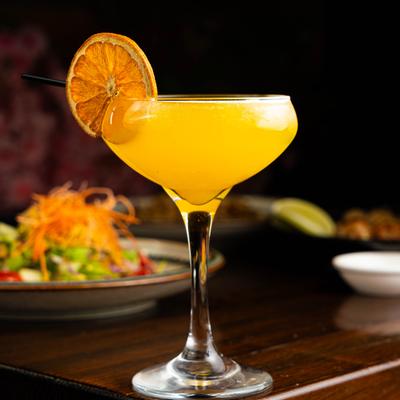 A vibrant orange cocktail garnished rests on a wooden table with food in the background.