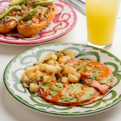Avocado tostadas with breakfast potatoes, and sandwiches on colorful plates beside a drink.