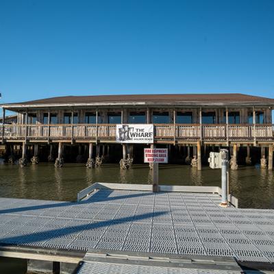 The Wharf exterior, A rustic wooden building on stilts, labeled  overlooks a calm waterfront.