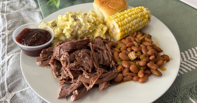Pulled pork plate with BBQ sauce, beans, corn, potato salad, and a dinner roll