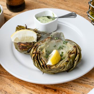 Grilled artichoke served with lemon wedges and a sauce on the side.