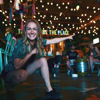 A smiling person making a hand gesture at a bar under string lights.