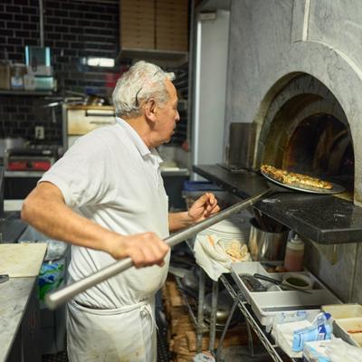 A pizza chef using a peel to tend to a pizza inside a wood-fired brick oven.