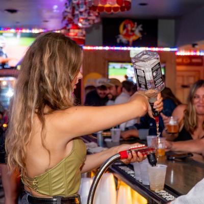 Bartender pours alcohol from a large bottle into a cup at a lively bar.