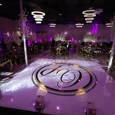 Elegant event hall with purple lighting, gold chairs, floral centerpieces, and a monogrammed white dance floor.