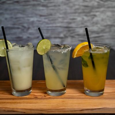 Three iced drinks on a wooden board, garnished with lime and orange slices..