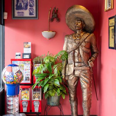 Life size bronze statue of a man with a sombrero beside a plant and a gumball machine.