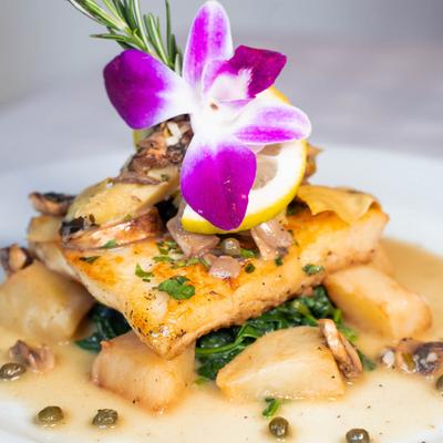 A close-up of grilled halibut topped with mushrooms, herbs, lemon, and an orchid.